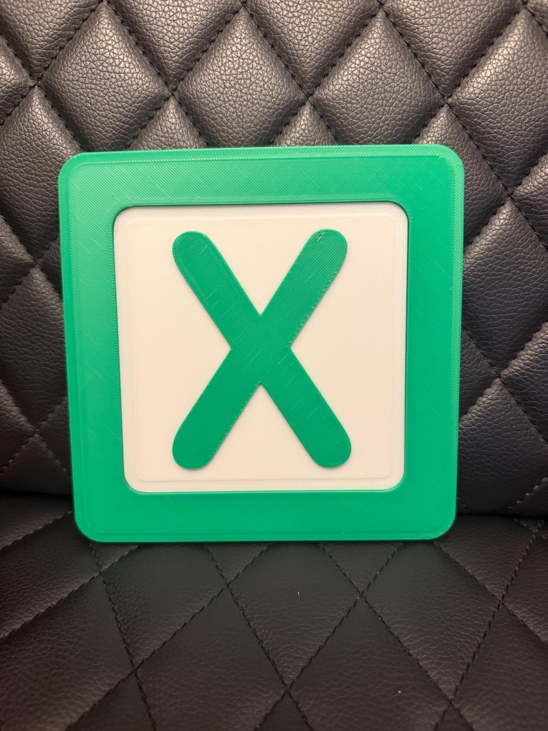 May include: A square, green-framed object with a white center featuring a large, green "X". The object is likely a game piece or a decorative item. The design is simple and bold, with a clear contrast between the green and white elements.