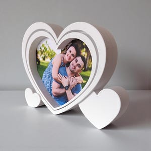 May include: A white heart-shaped photo frame with a double-layered design. The frame holds a colour photograph of a couple embracing. Two small heart-shaped supports are attached to the base of the frame.