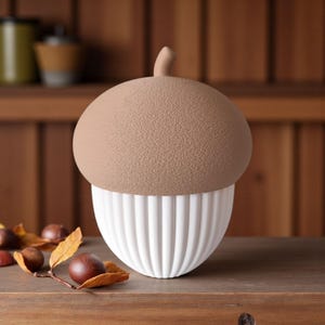 May include: A decorative acorn-shaped container. The top is a textured tan color, and the bottom is white with vertical ribbed detailing. The container has a small stem and sits on a wooden surface. Chestnuts and leaves are scattered around the base.