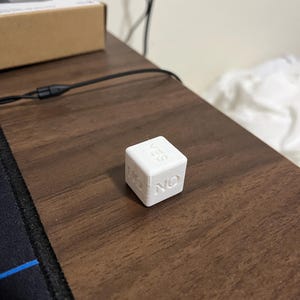 May include: A white decision die with the words "YES" and "NO" on adjacent sides. The die is sitting on a dark brown wooden surface. The die is a cube shape.