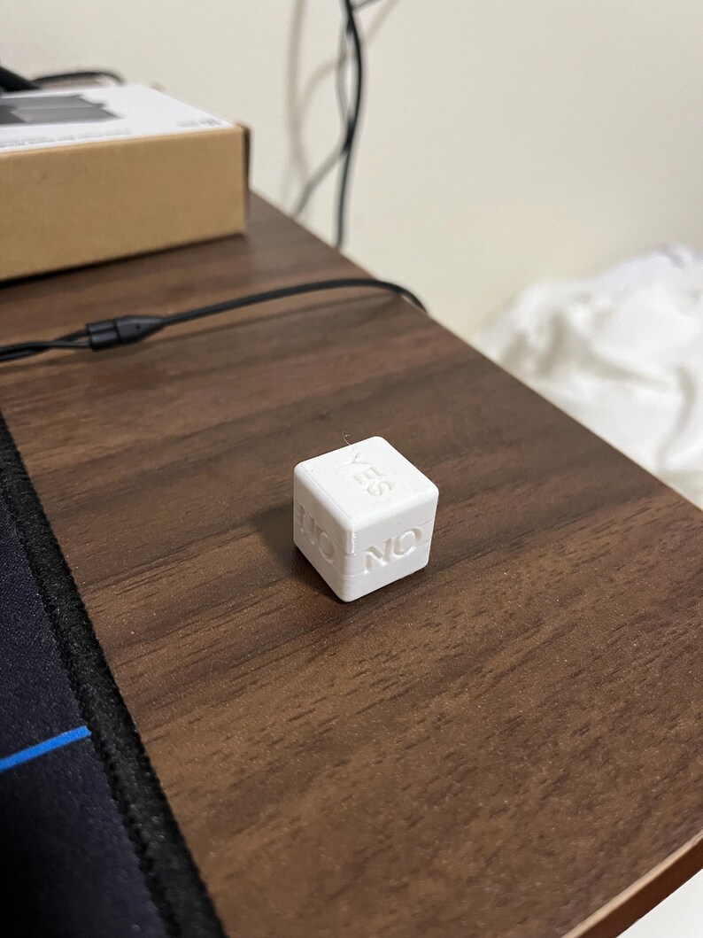 May include: A white decision die with the words "YES" and "NO" on adjacent sides. The die is sitting on a dark brown wooden surface. The die is a cube shape.