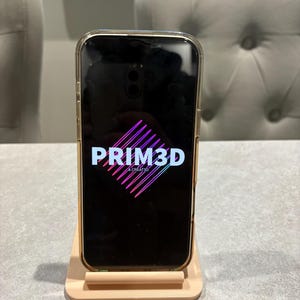 May include: A smartphone in a clear case displaying the PRIM3D logo in white and pink on a black screen. The phone is on a light beige phone stand. The logo is a stylized diamond shape with the text "& CREATED" below.