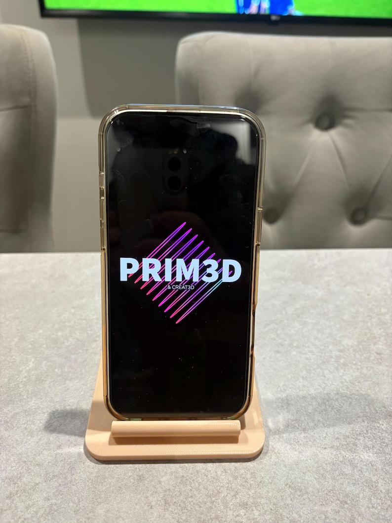 May include: A smartphone in a clear case displaying the PRIM3D logo in white and pink on a black screen. The phone is on a light beige phone stand. The logo is a stylized diamond shape with the text "& CREATED" below.