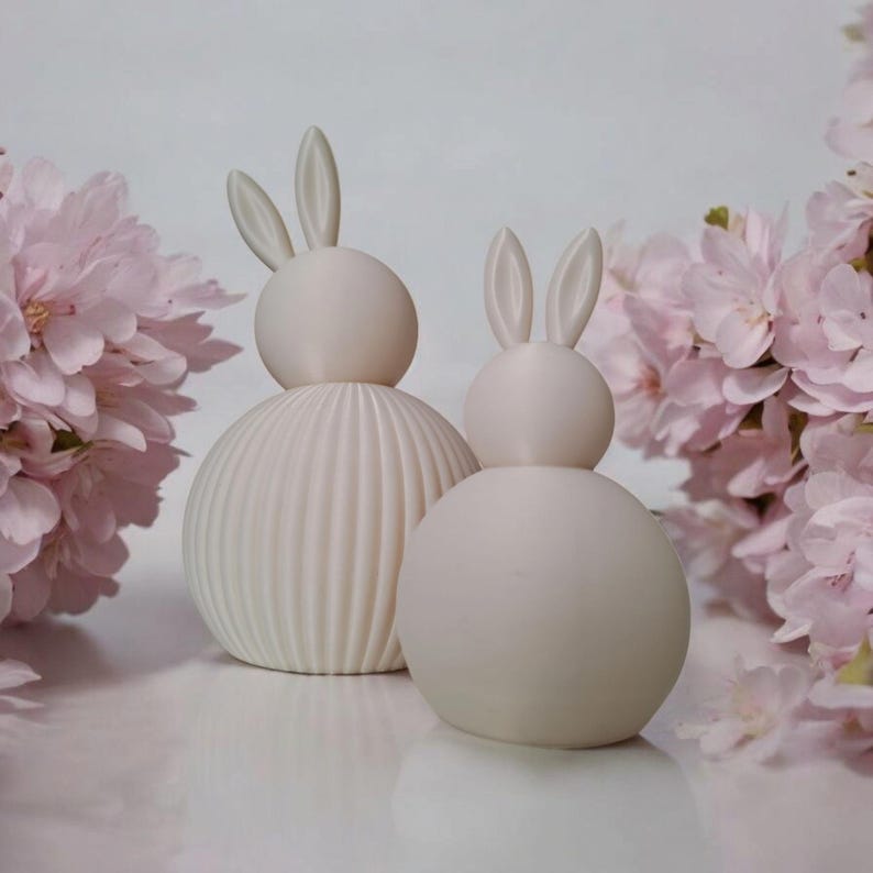May include: Two ceramic bunny figurines, one with a ribbed texture and the other smooth, both in a light beige color. The bunnies are sitting on a white surface with pink flowers in the background.