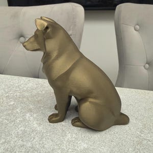 May include: A bronze-colored dog statue in a seated position, with its head turned to the left. The statue has a smooth finish and is placed on a light gray surface. The dog's ears are perked up, and its tail is tucked in.