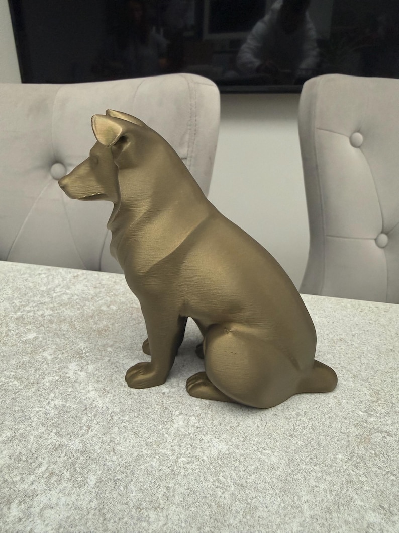 May include: A bronze-colored dog statue in a seated position, with its head turned to the left. The statue has a smooth finish and is placed on a light gray surface. The dog's ears are perked up, and its tail is tucked in.