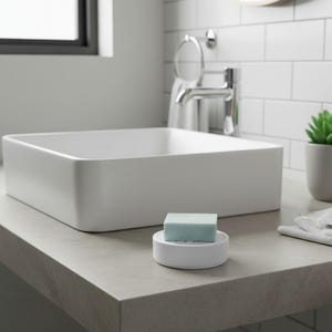 May include: A modern bathroom scene featuring a white, square sink and a light blue soap bar on a white soap dish. The sink sits on a gray countertop, with a window and faucet in the background. A small potted plant adds a touch of green.