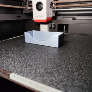 Pu&ograve; includere: Un vassoio rettangolare azzurro con un bordo anteriore curvo. Il vassoio &egrave; realizzato in un materiale solido e poggia su una superficie nera strutturata. Il vassoio &egrave; probabilmente stampato in 3D ed &egrave; un contenitore.