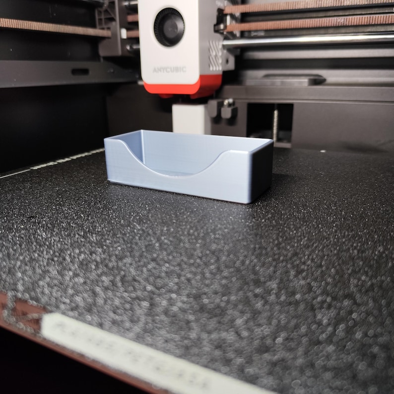 Pu&ograve; includere: Un vassoio rettangolare azzurro con un bordo anteriore curvo. Il vassoio &egrave; realizzato in un materiale solido e poggia su una superficie nera strutturata. Il vassoio &egrave; probabilmente stampato in 3D ed &egrave; un contenitore.