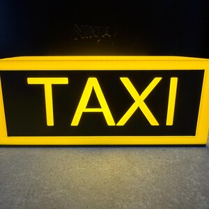 May include: A rectangular taxi sign with a bright yellow frame and the word "TAXI" in bold yellow letters against a black background. The sign is illuminated, suggesting it is lit from within. The sign is designed to be mounted on a vehicle.
