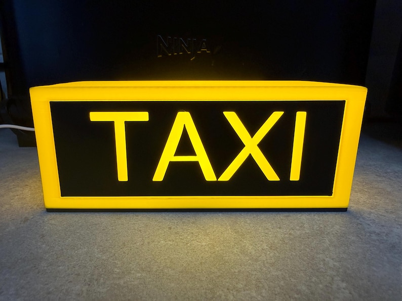 May include: A rectangular taxi sign with a bright yellow frame and the word "TAXI" in bold yellow letters against a black background. The sign is illuminated, suggesting it is lit from within. The sign is designed to be mounted on a vehicle.
