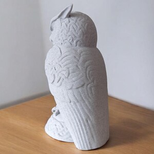 May include: A light gray owl figurine with detailed feather and wing patterns. The owl is perched on a small base and has a textured surface. The figurine is made of a speckled material and is displayed on a wooden surface.
