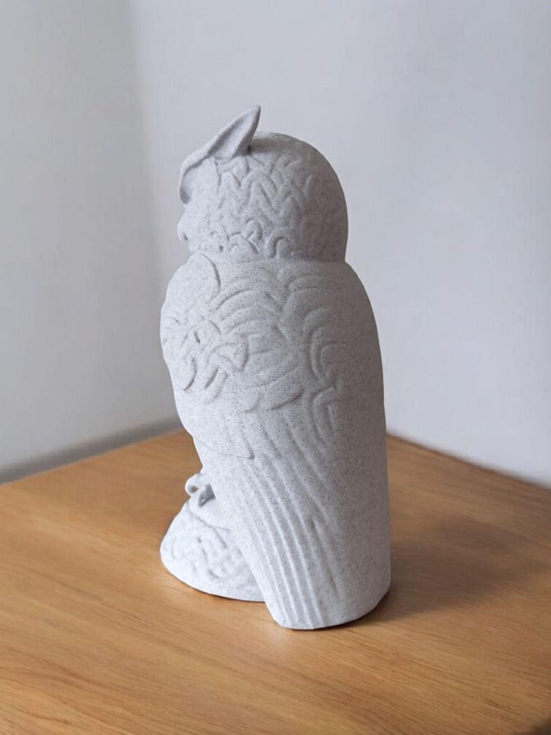 May include: A light gray owl figurine with detailed feather and wing patterns. The owl is perched on a small base and has a textured surface. The figurine is made of a speckled material and is displayed on a wooden surface.