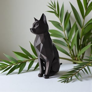 May include: A black geometric cat sculpture with a faceted design. The cat is sitting upright with its ears perked. Green leafy branches are in the background. The sculpture is on a white surface.