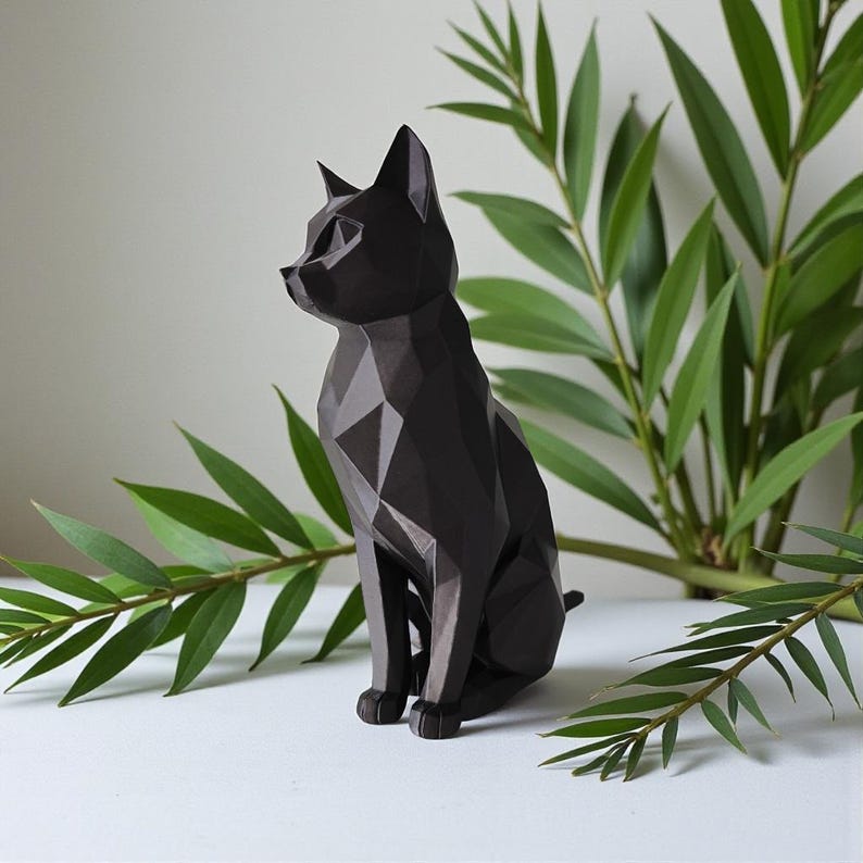 May include: A black geometric cat sculpture with a faceted design. The cat is sitting upright with its ears perked. Green leafy branches are in the background. The sculpture is on a white surface.