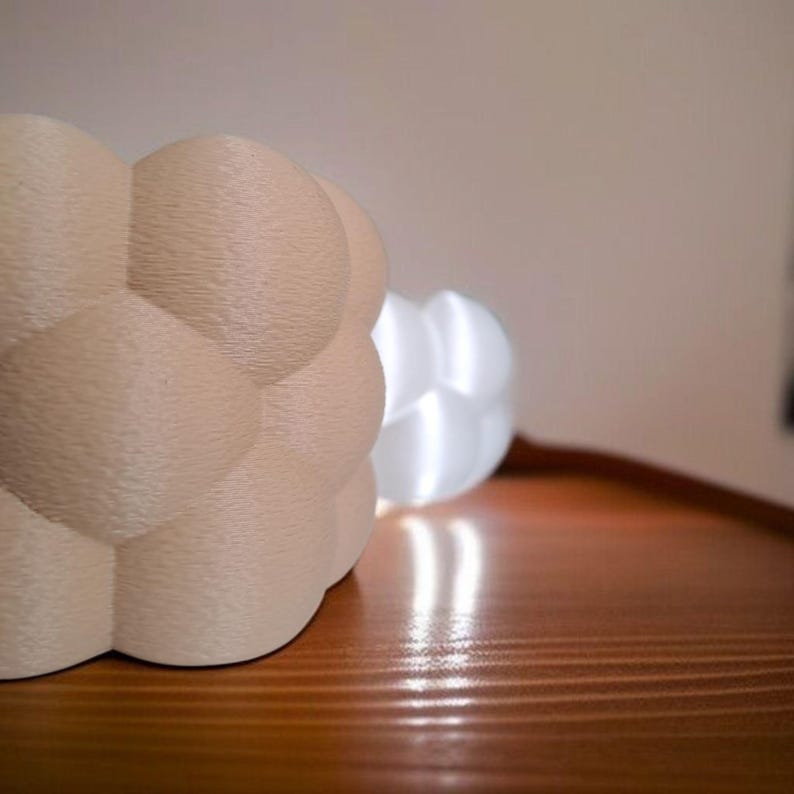 May include: Two decorative, spherical lamps with a textured, bubble-like design. One lamp is a light beige color, and the other is white. The lamps are sitting on a wooden surface, with the white lamp illuminated, casting a warm glow.