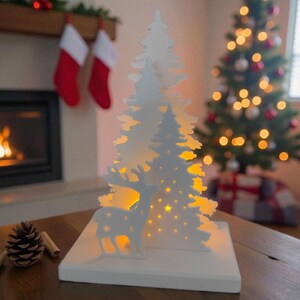 May include: A white Christmas-themed decorative light featuring a deer and a stylized tree design. The tree has star cutouts and is illuminated with warm yellow lights. The light sits on a white base, perfect for holiday decor.