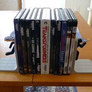 May include: A collection of Blu-ray movie cases, including "Transformers" and "Interstellar", held between two gray bookends with a running person design. The cases are stacked on a wooden table.