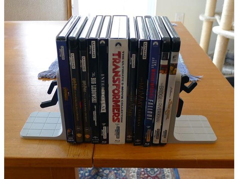 May include: A collection of Blu-ray movie cases, including "Transformers" and "Interstellar", held between two gray bookends with a running person design. The cases are stacked on a wooden table.
