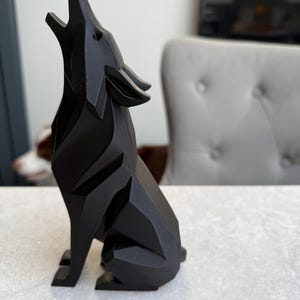 May include: A black, geometric wolf figurine in a howling pose. The sculpture has a faceted design, with sharp angles and a smooth, matte finish. The wolf is seated on its hind legs, with its head and snout raised towards the sky.