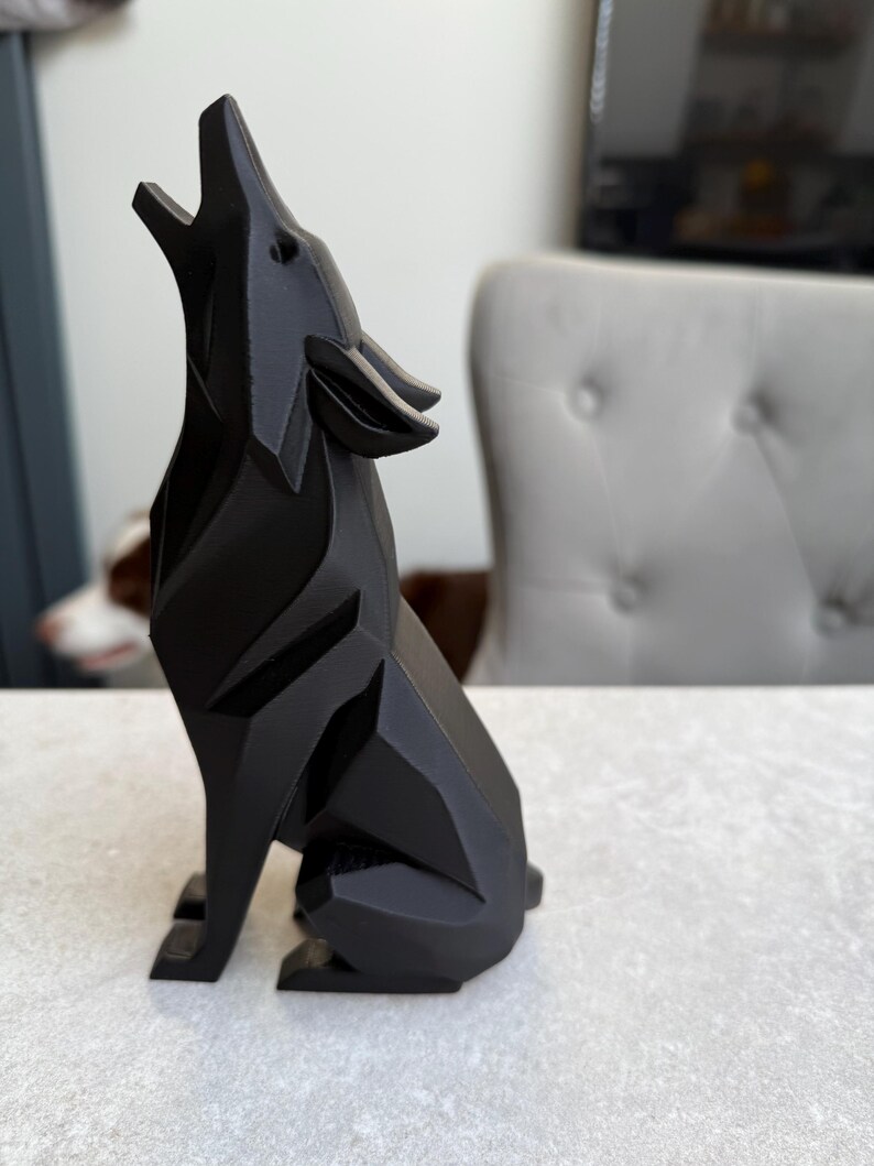 May include: A black, geometric wolf figurine in a howling pose. The sculpture has a faceted design, with sharp angles and a smooth, matte finish. The wolf is seated on its hind legs, with its head and snout raised towards the sky.