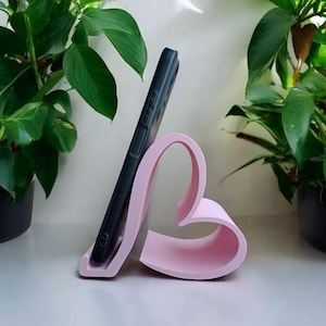 May include: A pink heart-shaped phone stand with a black smartphone resting in it. The stand is on a white surface with two green plants in pots on either side.