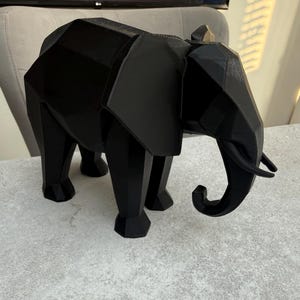 Op de afbeelding: Een zwarte geometrische olifant sculptuur. Het decoratieve stuk heeft een polygonale vormgeving met zichtbare randen en een omhoog gekrulde slurf. De olifant staat op vier poten en heeft grote oren. De sculptuur is gemaakt van een effen, matzwart materiaal.