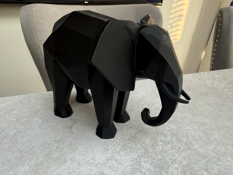 Op de afbeelding: Een zwarte geometrische olifant sculptuur. Het decoratieve stuk heeft een polygonale vormgeving met zichtbare randen en een omhoog gekrulde slurf. De olifant staat op vier poten en heeft grote oren. De sculptuur is gemaakt van een effen, matzwart materiaal.
