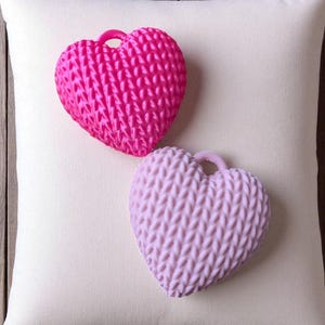 May include: Two heart-shaped decorative objects with a textured knit pattern, one bright pink and the other light purple, rest on a white pillow. The hearts have a small loop at the top. The background is a wooden surface.