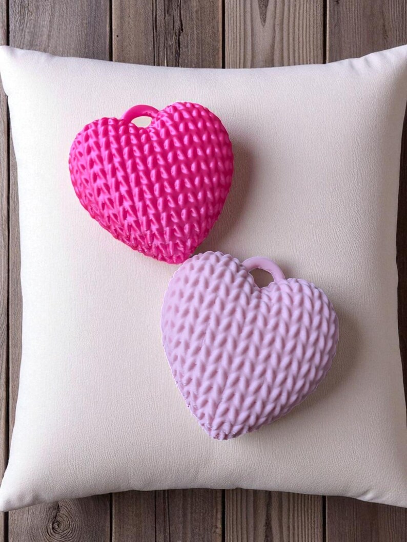 May include: Two heart-shaped decorative objects with a textured knit pattern, one bright pink and the other light purple, rest on a white pillow. The hearts have a small loop at the top. The background is a wooden surface.