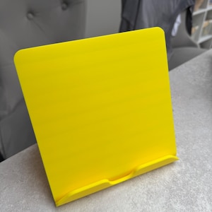 May include: A bright yellow book stand made of plastic. The stand is rectangular with a curved top and a small ledge at the bottom to hold a book or tablet. The stand is on a gray surface.