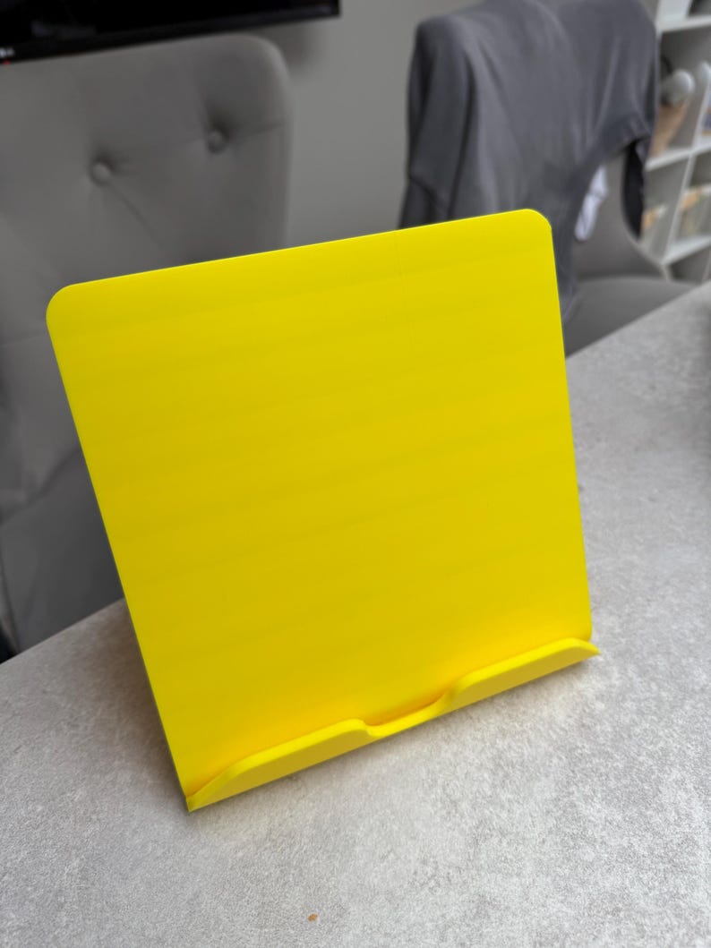May include: A bright yellow book stand made of plastic. The stand is rectangular with a curved top and a small ledge at the bottom to hold a book or tablet. The stand is on a gray surface.