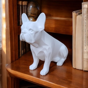 May include: White ceramic figurine of a French bulldog sitting on a wooden shelf. The figurine is detailed and realistic, with a smooth, matte finish.