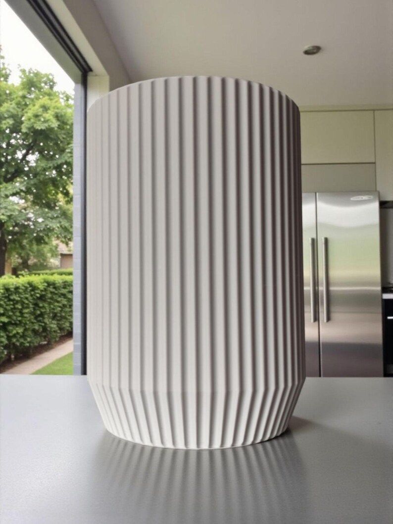 May include: A tall, cylindrical, off-white vase with vertical ribbed detailing. The vase has a slightly flared base and stands on a gray surface. The background includes a window with a view of greenery and a modern kitchen.