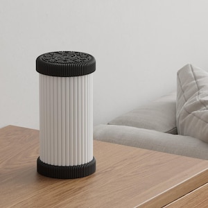 Pode incluir: L&acirc;mpada cil&iacute;ndrica branca e preta com parte superior e inferior texturizadas e linhas verticais no corpo. A l&acirc;mpada est&aacute; sobre uma mesa de madeira, com um sof&aacute; ao fundo. Decora&ccedil;&atilde;o de casa.