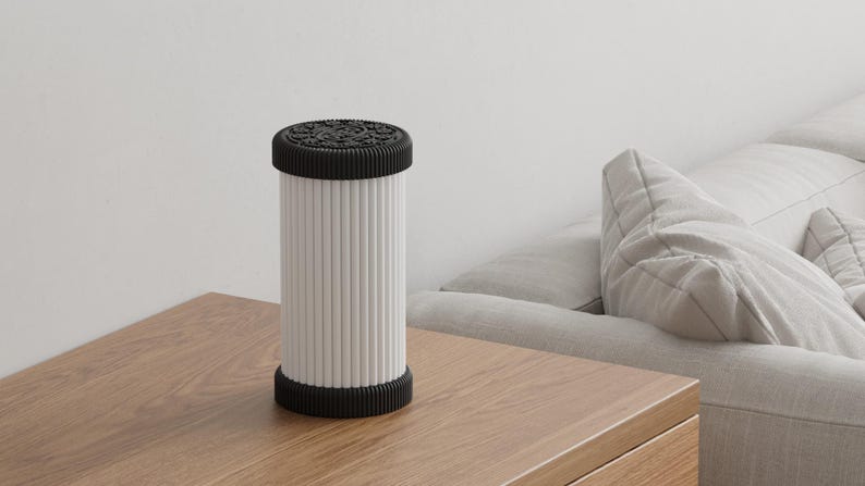 Pode incluir: L&acirc;mpada cil&iacute;ndrica branca e preta com parte superior e inferior texturizadas e linhas verticais no corpo. A l&acirc;mpada est&aacute; sobre uma mesa de madeira, com um sof&aacute; ao fundo. Decora&ccedil;&atilde;o de casa.