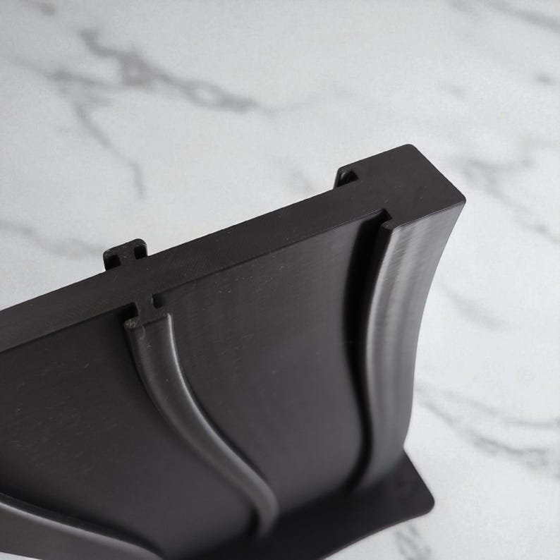 May include: A close-up of a black plastic object with a curved design. The object has a rectangular top with two small, raised tabs. The curved portion has a smooth, flowing shape. The background is a blurred, light-coloured surface.
