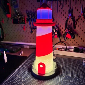 May include: A decorative lighthouse lamp with a red and white striped design. The lamp features a red top with a blue light, a red door, and a black base. The lamp is approximately 12 inches tall.