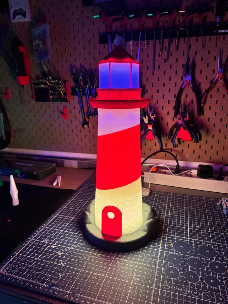 May include: A decorative lighthouse lamp with a red and white striped design. The lamp features a red top with a blue light, a red door, and a black base. The lamp is approximately 12 inches tall.