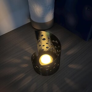 May include: A black metal candle holder with a semi-circular design and numerous circular cutouts. A lit, round candle is inside, casting a warm glow. The holder sits on a wooden surface, with a white ceramic object in the background.