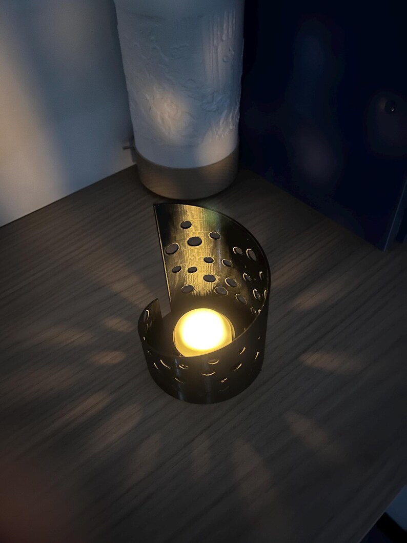 May include: A black metal candle holder with a semi-circular design and numerous circular cutouts. A lit, round candle is inside, casting a warm glow. The holder sits on a wooden surface, with a white ceramic object in the background.