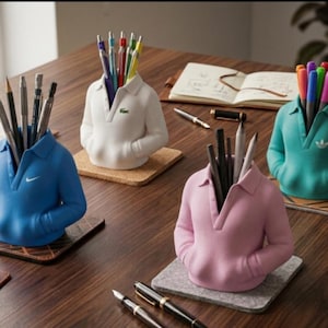 Puede incluir: Cuatro portal&aacute;pices con forma de polos en azul, blanco, rosa y turquesa, cada uno con bol&iacute;grafos y l&aacute;pices. Los soportes est&aacute;n sobre posavasos en una mesa de madera. Los logotipos de Nike, Lacoste y Adidas son visibles.
