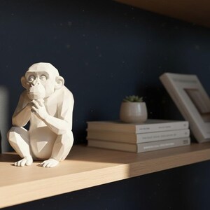 May include: A white, geometric monkey sculpture sits on a wooden shelf. The sculpture is in a seated position with its hands clasped. Other items on the shelf include a small potted plant, and several books. The background is a dark blue wall.