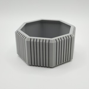 Puede incluir: Una maceta hexagonal gris con detalles acanalados verticales en los lados. El interior es de un tono gris m&aacute;s oscuro. La maceta est&aacute; dise&ntilde;ada con una est&eacute;tica moderna, adecuada para uso en interiores o exteriores.