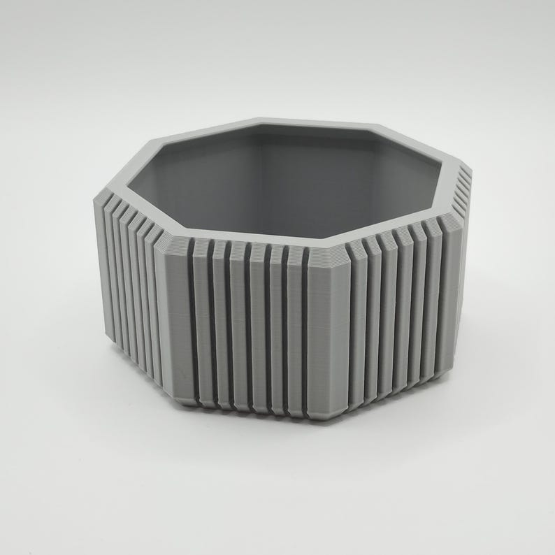 Puede incluir: Una maceta hexagonal gris con detalles acanalados verticales en los lados. El interior es de un tono gris m&aacute;s oscuro. La maceta est&aacute; dise&ntilde;ada con una est&eacute;tica moderna, adecuada para uso en interiores o exteriores.
