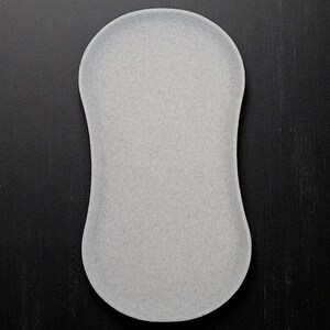 May include: A light grey, oval-shaped tray with a speckled texture. The tray has rounded edges and a smooth surface. The background is a dark, textured surface.