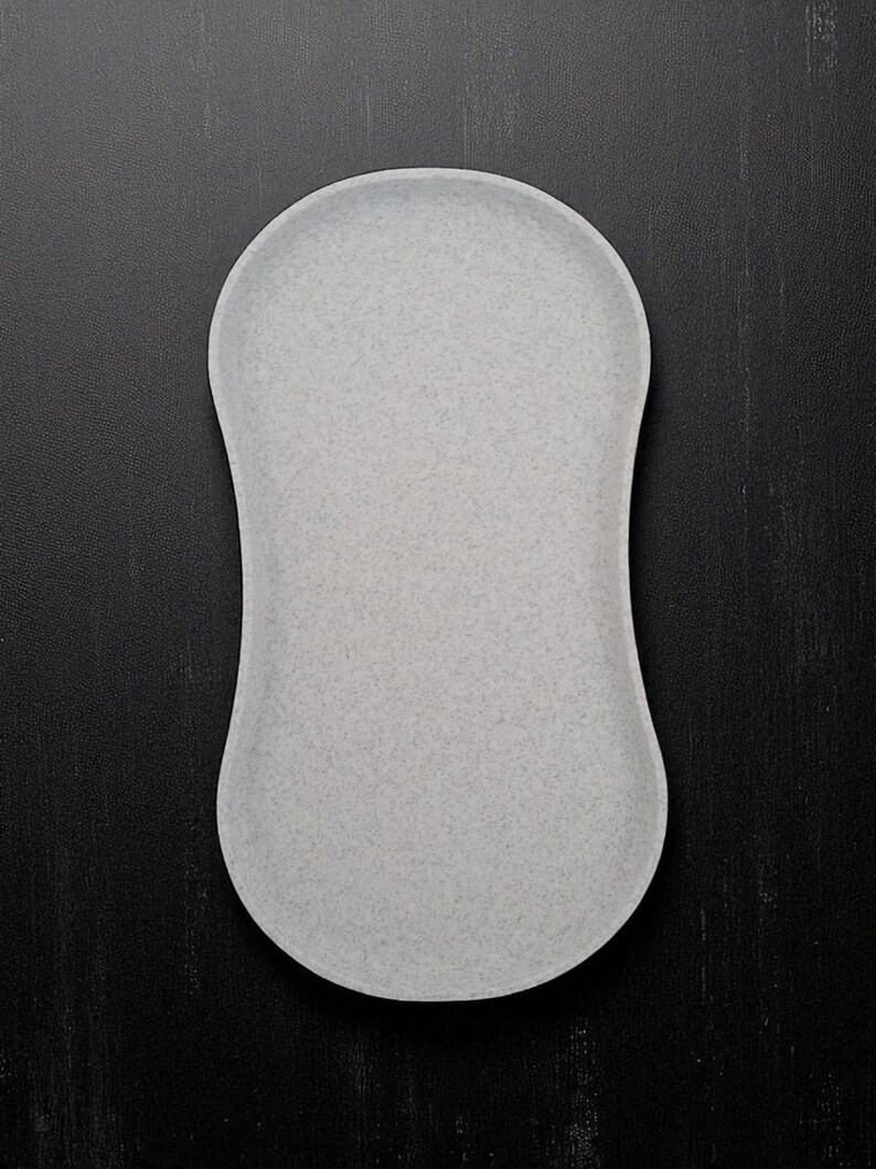 May include: A light grey, oval-shaped tray with a speckled texture. The tray has rounded edges and a smooth surface. The background is a dark, textured surface.