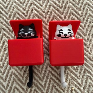 May include: Two red cat-shaped hooks. One hook features a black cat, the other a white cat. The cats are inside red boxes with the lids open. The hooks are black and white, and the background is a beige and brown zigzag pattern.