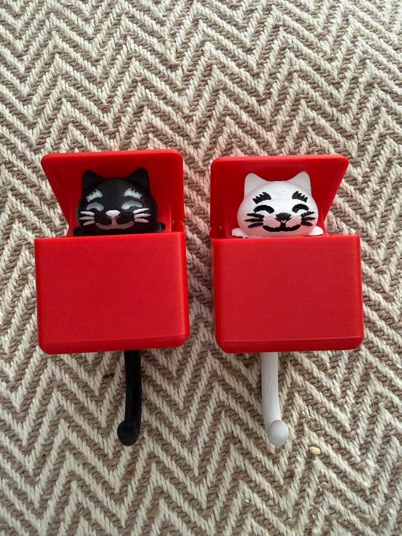 May include: Two red cat-shaped hooks. One hook features a black cat, the other a white cat. The cats are inside red boxes with the lids open. The hooks are black and white, and the background is a beige and brown zigzag pattern.