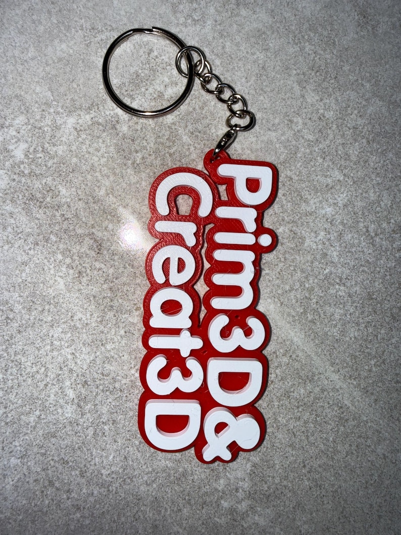 May include: A red and white keychain with the text "Prim3D & Create3D". The keychain has a silver keyring and chain. The text is in a bold, sans-serif font.